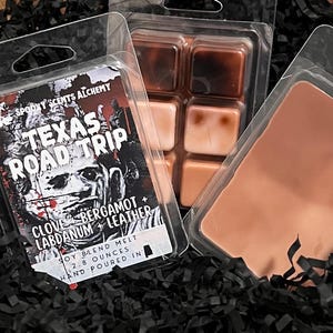 May include: Wax melts in clear plastic packaging. One package features the text "TEXAS ROAD TRIP" with "CLOVE BERGAMOT + LEATHER" and "SOY BLEND MELT 2.8 OUNCES HAND POURED IN". Other packages contain brown and tan wax cubes.