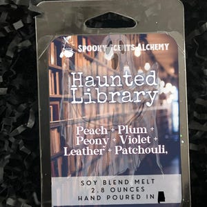 May include: A soy blend melt in a clear plastic package. The label reads "Haunted Library" with scent notes of peach, plum, peony, violet, leather, and patchouli. It weighs 2.8 ounces and is hand-poured.