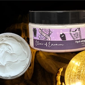 May include: A white hydrating mineral mask in a purple and black jar with a spooky design. The jar has the text "Elixir of Evermore" and "Hydrating Mineral Mask" on it. The jar is sitting on a gold surface.