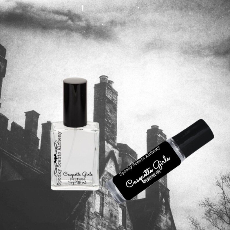 May include: Two bottles of perfume oil, one in a clear glass bottle with a black cap and the other in a rollerball bottle. Both bottles have black labels with white text that reads "Spooky Scents Alchemy" and "Casquette Girls". The larger bottle is labeled "Perfume 1 fl oz/30 ml". The smaller bottle is labeled "Perfume Oil".
