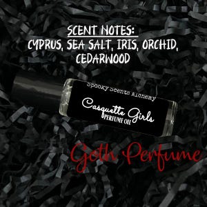 May include: A black roll-on bottle with a white label that reads "Spooky Scents Alchemy Casquette Girls Perfume Oil". The scent notes are listed as "Cyprus, Sea Salt, Iris, Orchid, Cedarwood".