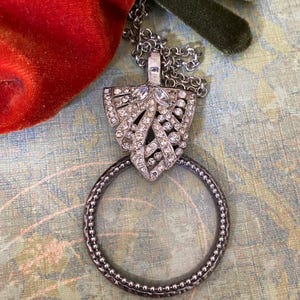 May include: A silver-toned magnifying glass pendant necklace. The pendant features a decorative shield shape adorned with clear crystals. The circular magnifying glass has a beaded edge and hangs from a delicate chain.