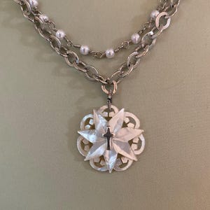 Può includere: Una collana color argento con due fili. Il filo superiore presenta piccole perle bianche, mentre il filo inferiore ha una catena spessa. Un ciondolo con un design a forma di fiore in madreperla e una piccola croce pende dalla catena.