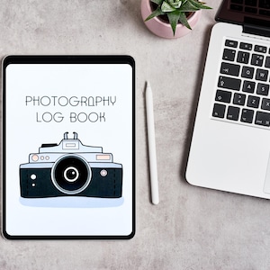 May include: A white digital tablet with a black camera icon and the text "PHOTOGRAPHY LOG BOOK" on the screen. The tablet is on a gray surface with a white stylus and a laptop computer.