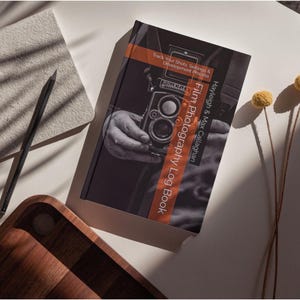 Op de afbeelding: Een bruin boek met de titel "Film Photography Log Book" van Hayleigh & Max Callaghan. De omslag toont een foto van een hand die een vintage filmcamera vasthoudt.