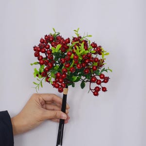 Może przedstawiać: Bukiet sztucznych jagód z małymi, okrągłymi, ciemnoczerwonymi owocami i jasnozielonymi liśćmi. Jagody są zgrupowane, a łodygi są ciemnobrązowe. Bukiet jest trzymany na gładkim, białym tle.