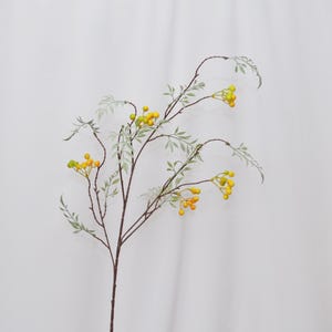 Może przedstawiać: Dekoracyjna łodyga sztucznej rośliny z cienkimi, brązowymi gałązkami i małymi, jasnozielonymi liśćmi. Skupiska żółtych jagód są rozrzucone wzdłuż łodyg, tworząc naturalną, botaniczną estetykę. Idealna do kompozycji kwiatowych i dekoracji wnętrz.