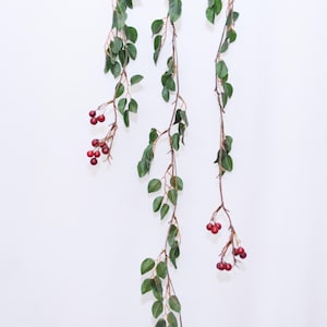 May include: Three strands of artificial greenery hang against a white backdrop. Each strand features small, oval green leaves and clusters of small, round, red berries. The stems are brown and slightly textured.