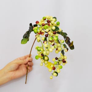 Może przedstawiać: Gałązka sztucznych jagód w różnych kolorach, w tym żółtym, zielonym i kasztanowym. Jagody są zgrupowane i przymocowane do brązowej łodygi z zielonymi liśćmi. Gałązka jest trzymana przez dłoń na białym tle.