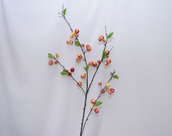 May include: A branch with small green leaves and red and yellow artificial apples.