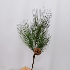 May include: A faux pine branch with long, green needles and a small brown pinecone. The branch has a dark brown stem and is set against a plain white background. This artificial plant is suitable for seasonal decor.