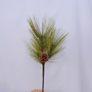 May include: An artificial pine branch featuring green needles and a brown pinecone. The branch has a dark brown stem, set against a white backdrop. This decorative item is suitable for seasonal home decor.