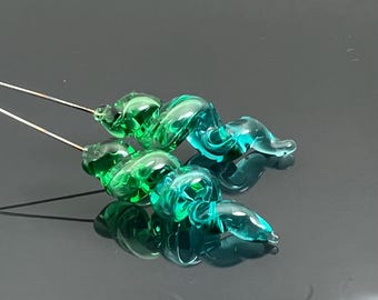 Könnte beinhalten: Zwei Glasperlen mit einem wirbelnden, marmorierten Design in Grün- und Blautönen. Die Perlen sind an dünnen Metalldrähten befestigt.