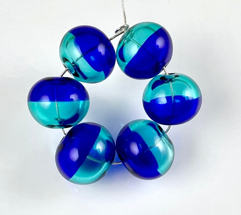May include: A circular arrangement of six glass beads. Each bead is a sphere, split diagonally into two colors: a vibrant blue and a teal. The beads are strung together with thin, silver-colored wire, creating a decorative element.