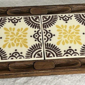 Peut inclure: Un dessous de plat rectangulaire en bois avec deux carreaux de céramique. Les carreaux ont un fond crème avec des motifs floraux jaunes et bruns. Le cadre en bois est de couleur marron foncé et comporte des détails sculptés.