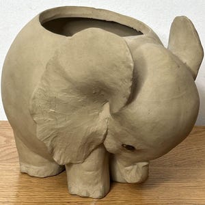 May include: A beige ceramic elephant planter with a large opening at the top. The elephant has large ears, a trunk, and four legs. The planter is designed to hold plants or flowers.