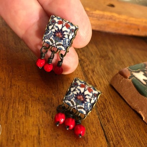 Puede incluir: Un par de pendientes con un colgante de forma cuadrada con un patrón floral azul y blanco. El colgante está unido a una cadena colgante con tres cuentas rojas.