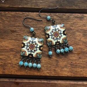 May include: A pair of square dangle earrings with a floral tile design in cream, brown, and blue. Each earring has a fringe of small blue beads. The earrings are displayed on a wooden surface with a branch.