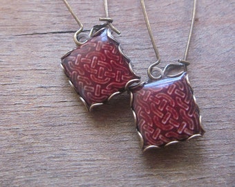 May include: A pair of handcrafted earrings with a square, burgundy-colored enamel design. The earrings have a gold-colored metal frame and long, curved ear wires. The square design features a repeating pattern.