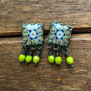 Pu&ograve; includere: Un paio di orecchini quadrati con un motivo floreale. Gli orecchini presentano uno sfondo verde chiaro con fiori blu e bianchi. Ogni orecchino ha tre perline pendenti verde neon. Gli orecchini sono appoggiati su una superficie di legno.