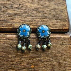 May include: A pair of floral stud earrings. Each earring features a blue flower design with green accents, set in a round, antique-style metal frame. Dangling below are three small, striped beads. The earrings rest on a wooden surface.