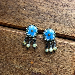 May include: A pair of floral stud earrings with blue and white flower designs. Each earring has three dangling strands with small, round, cream-colored beads. The earrings are set against a wooden background, with the text "CORINA CROOKS" in a circle.