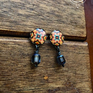 May include: A pair of earrings with a circular, patterned top in red, orange, and yellow, with a black metal frame. The earrings dangle with a dark blue, rectangular bead. The earrings are displayed on a wooden surface. The text "CORINA CROOKS" is in a white circle.