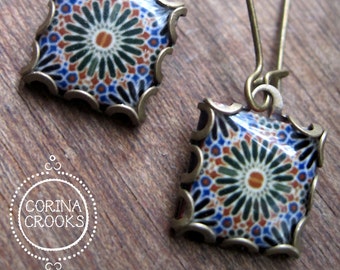 Può includere: Un paio di orecchini pendenti quadrati con un design a mosaico. Gli orecchini presentano una montatura color ottone e un colorato motivo a mosaico con dettagli blu, arancioni e verdi. Sono appoggiati su una superficie di legno.