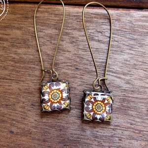 May include: A pair of earrings with a square, floral design in shades of yellow, brown, and white. The earrings have long, gold-colored hooks. The brand name "CORINA CROOKS" is in a circle in the upper left corner.
