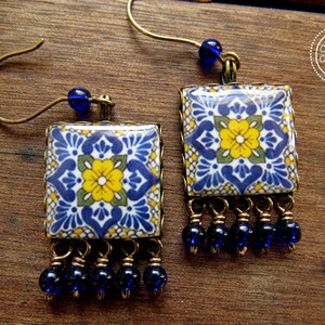 May include: A pair of handcrafted earrings featuring square ceramic tiles with a blue, yellow, and green floral pattern. The earrings have brass-colored metal frames and are adorned with dangling blue beads. The earrings are designed with a hook closure.