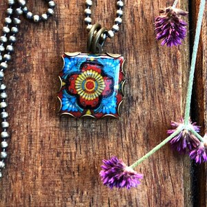 May include: A square pendant necklace with a colorful floral design in red, yellow, and blue, set in a bronze-colored frame. The necklace hangs on a silver ball chain. A sprig of purple flowers rests on a wooden surface. The text "CORINA CROOKS" is visible.