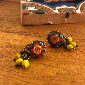 May include: A pair of earrings with a floral design. The earrings have a dark metal base with a red and yellow floral pattern. Each earring has a cluster of yellow beads hanging from it. The earrings are on a wooden surface.