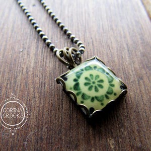 May include: A green and white ceramic tile pendant necklace with a brass chain. The tile has a floral design.