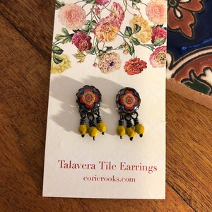 May include: A pair of Talavera tile earrings with a floral design. The earrings have a dark blue background with a red and yellow floral pattern. Each earring has three yellow beads dangling from the bottom. The earrings are displayed on a white card with the text "Talavera Tile Earrings" and the website coricrooks.com.