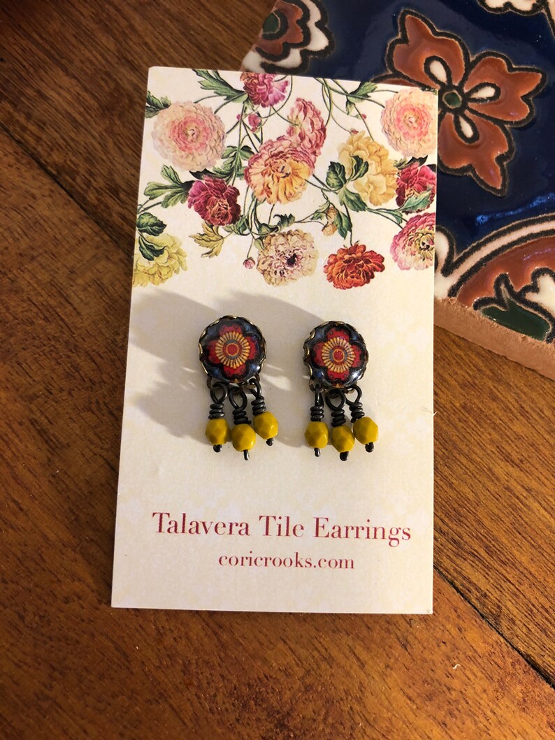 May include: A pair of Talavera tile earrings with a floral design. The earrings have a dark blue background with a red and yellow floral pattern. Each earring has three yellow beads dangling from the bottom. The earrings are displayed on a white card with the text "Talavera Tile Earrings" and the website coricrooks.com.