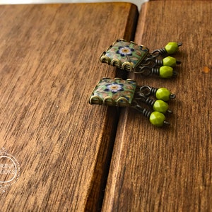 Pu&ograve; includere: Un paio di orecchini quadrati con un design floreale. Gli orecchini hanno un motivo a mosaico con colori verdi, blu e bianchi. Ogni orecchino ha tre perline verdi appese. Gli orecchini sono appoggiati su una superficie di legno.