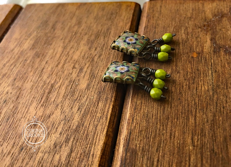 Pu&ograve; includere: Un paio di orecchini quadrati con un design floreale. Gli orecchini hanno un motivo a mosaico con colori verdi, blu e bianchi. Ogni orecchino ha tre perline verdi appese. Gli orecchini sono appoggiati su una superficie di legno.