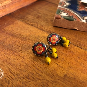 May include: A pair of handcrafted earrings with a floral design. The earrings feature a dark blue background with a red and yellow flower pattern. Dangling from the earrings are yellow beads. The earrings are displayed on a wooden surface.