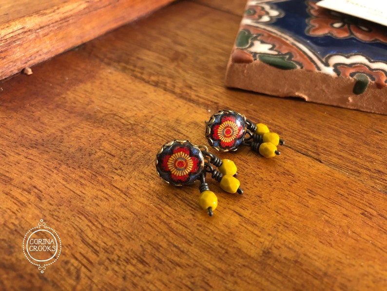May include: A pair of handcrafted earrings with a floral design. The earrings feature a dark blue background with a red and yellow flower pattern. Dangling from the earrings are yellow beads. The earrings are displayed on a wooden surface.
