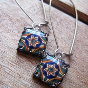 Puede incluir: Un par de pendientes colgantes cuadrados con un diseño floral. Los pendientes presentan un patrón floral azul, naranja y verde dentro de un marco decorativo. Los ganchos para las orejas son de color dorado claro.