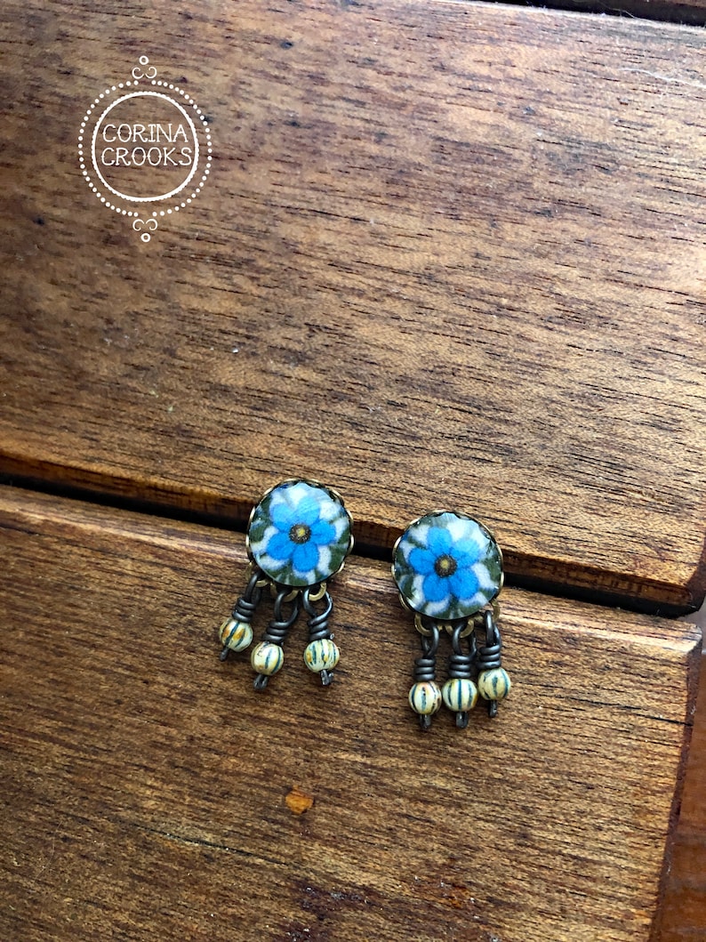 May include: A pair of floral stud earrings with blue and white flower designs. Each earring has three dangling strands with small, round, cream-colored beads. The earrings are set against a wooden background, with the text "CORINA CROOKS" in a circle.