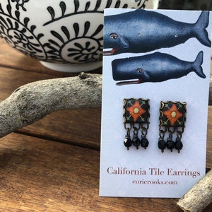 May include: A pair of square earrings with a floral design in orange and white on a black background. The earrings have a bronze-colored frame and dangle with three black beads. The card behind the earrings reads "California Tile Earrings".