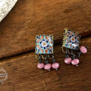 May include: A pair of square earrings with a blue and white floral design on the front. The earrings have a gold-colored frame and dangle pink beads.