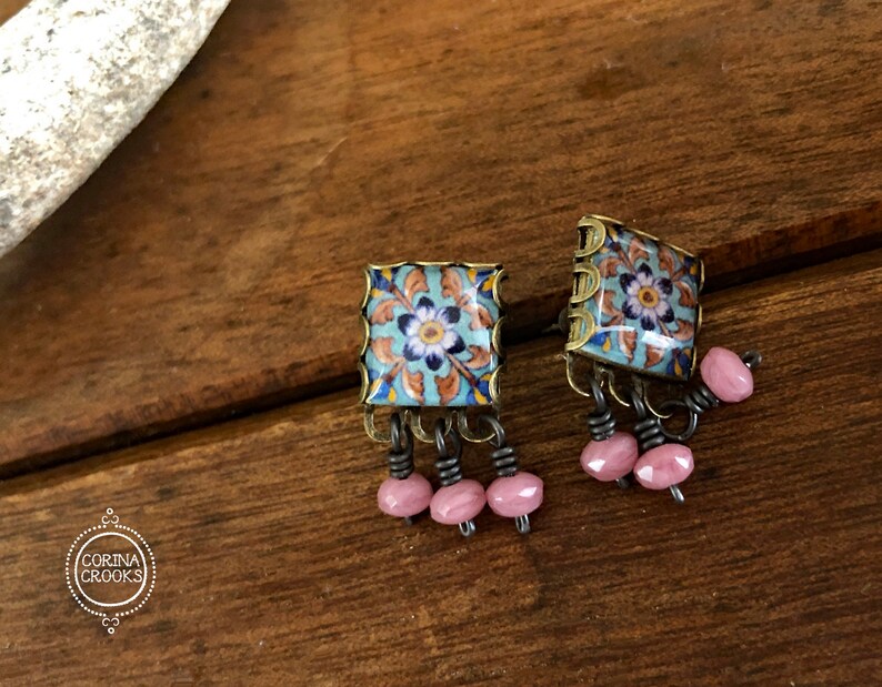 May include: A pair of square earrings with a blue and white floral design on the front. The earrings have a gold-colored frame and dangle pink beads.