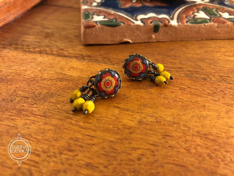 May include: A pair of earrings with a floral design. The earrings have a dark metal base with a red and yellow floral pattern. Each earring has a cluster of yellow beads hanging from it. The earrings are on a wooden surface.