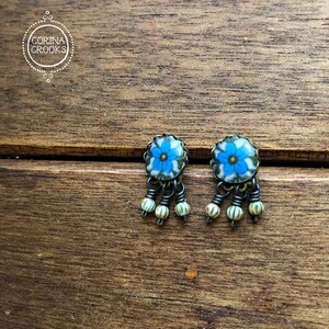 May include: A pair of dangle earrings featuring a blue floral design. The earrings have a silver-tone metal frame and are adorned with small, cream-colored beads. The earrings are displayed on a wooden surface. The brand name "CORINA CROOKS" is in a circle in the upper left corner.