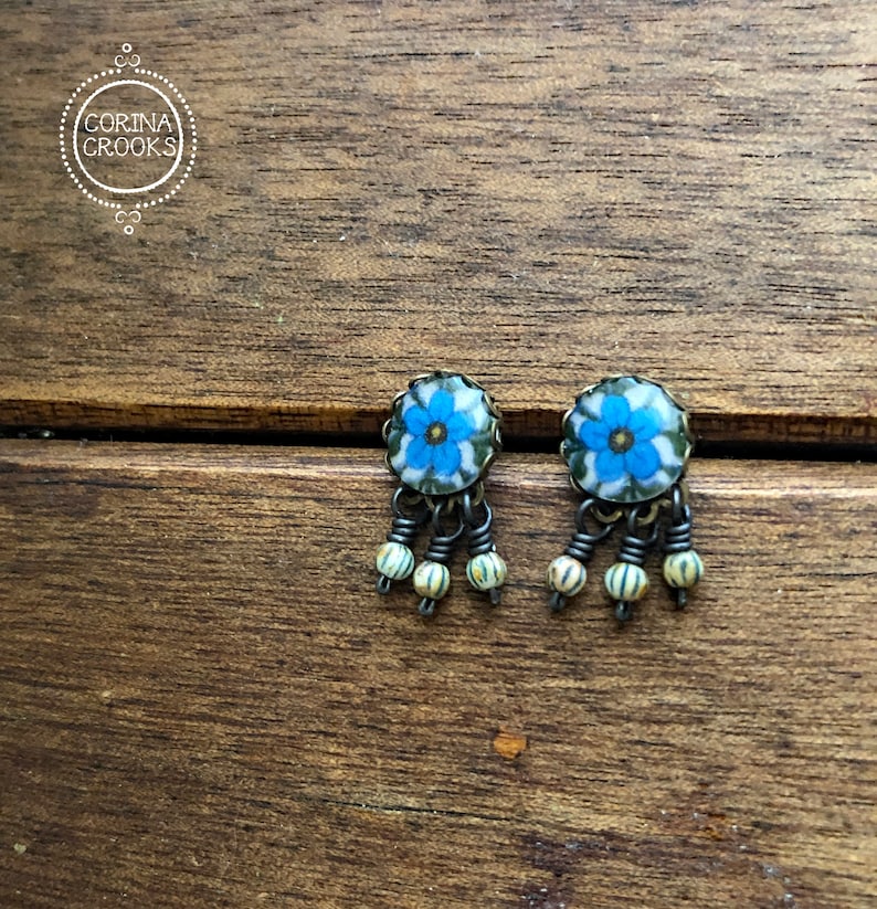 May include: A pair of dangle earrings featuring a blue floral design. The earrings have a silver-tone metal frame and are adorned with small, cream-colored beads. The earrings are displayed on a wooden surface. The brand name "CORINA CROOKS" is in a circle in the upper left corner.