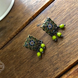 Pu&ograve; includere: Un paio di orecchini quadrati con un motivo floreale verde, blu e bianco. Gli orecchini hanno una montatura color bronzo e sono adornati da tre perline verde lime. Sono esposti su una superficie di legno.