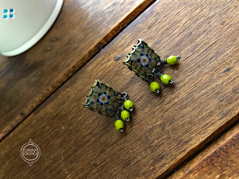 Pu&ograve; includere: Un paio di orecchini quadrati con un motivo floreale verde, blu e bianco. Gli orecchini hanno una montatura color bronzo e sono adornati da tre perline verde lime. Sono esposti su una superficie di legno.