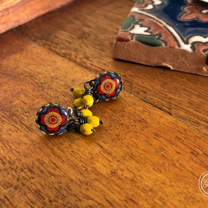 May include: Close-up of handmade earrings with a floral design. The earrings feature a red and yellow flower pattern set in a dark blue frame. Yellow beads dangle below the floral design. The earrings are on a wooden surface.